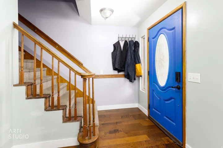 Welcoming foyer featuring a striking cobalt blue entry door with decorative glass panel, warm wood-finish hardwood flooring, and a wooden staircase with turned balusters and handrail