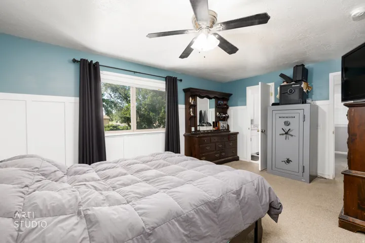 Spacious primary bedroom showcasing soft blue accent walls, ceiling fan with integrated lighting, large multi-pane windows with blackout curtains, neutral carpet flooring, and ample closet storage