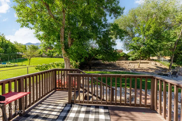 Wood-tone deck with a slatted railing overlooking a manicured lawn, mature trees, and landscaping elements