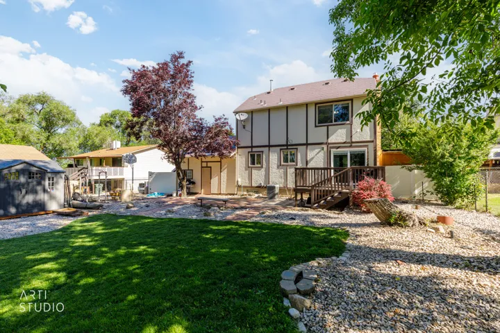 Spacious backyard oasis featuring a manicured lawn, mature landscaping with flowering shrubs, gravel hardscaping, and a two-story Colonial-style home with wood and stone exterior, rear deck, and detached outbuildings