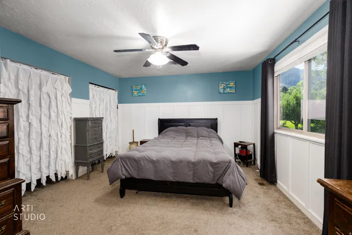 Spacious primary bedroom with two-tone paint featuring turquoise upper walls and white wainscoting, ceiling fan with integrated lighting, carpet flooring, and large sliding glass doors with dark frame opening to exterior grounds
