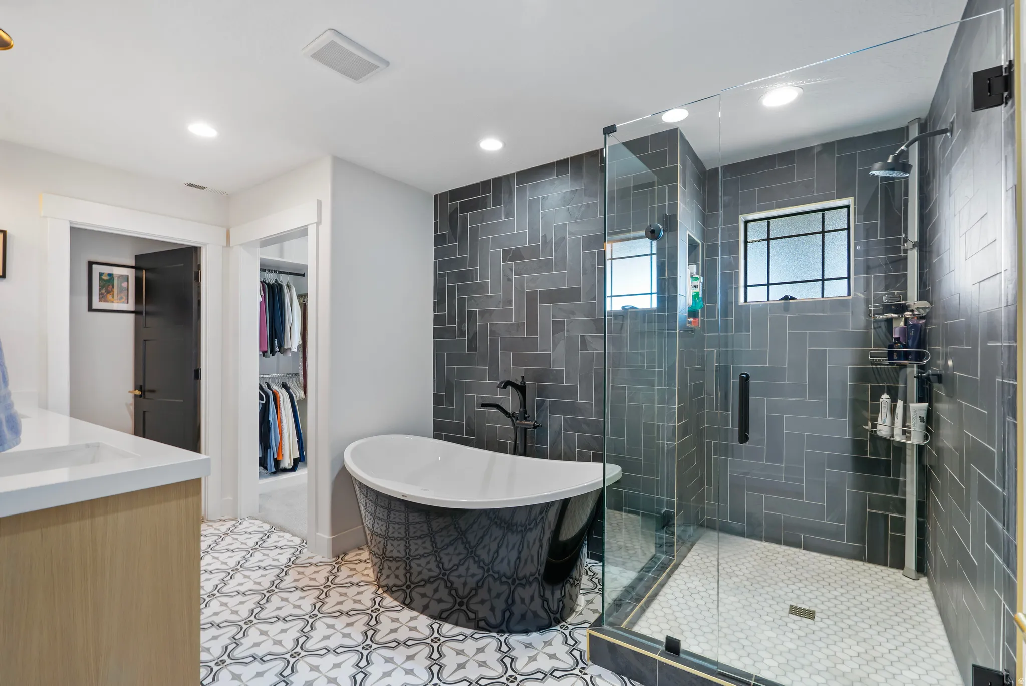 Contemporary bathroom featuring a patterned freestanding tub, a glass-enclosed shower with herringbone tile, a single vanity with a white countertop, patterned floor tile, and a walk-in closet