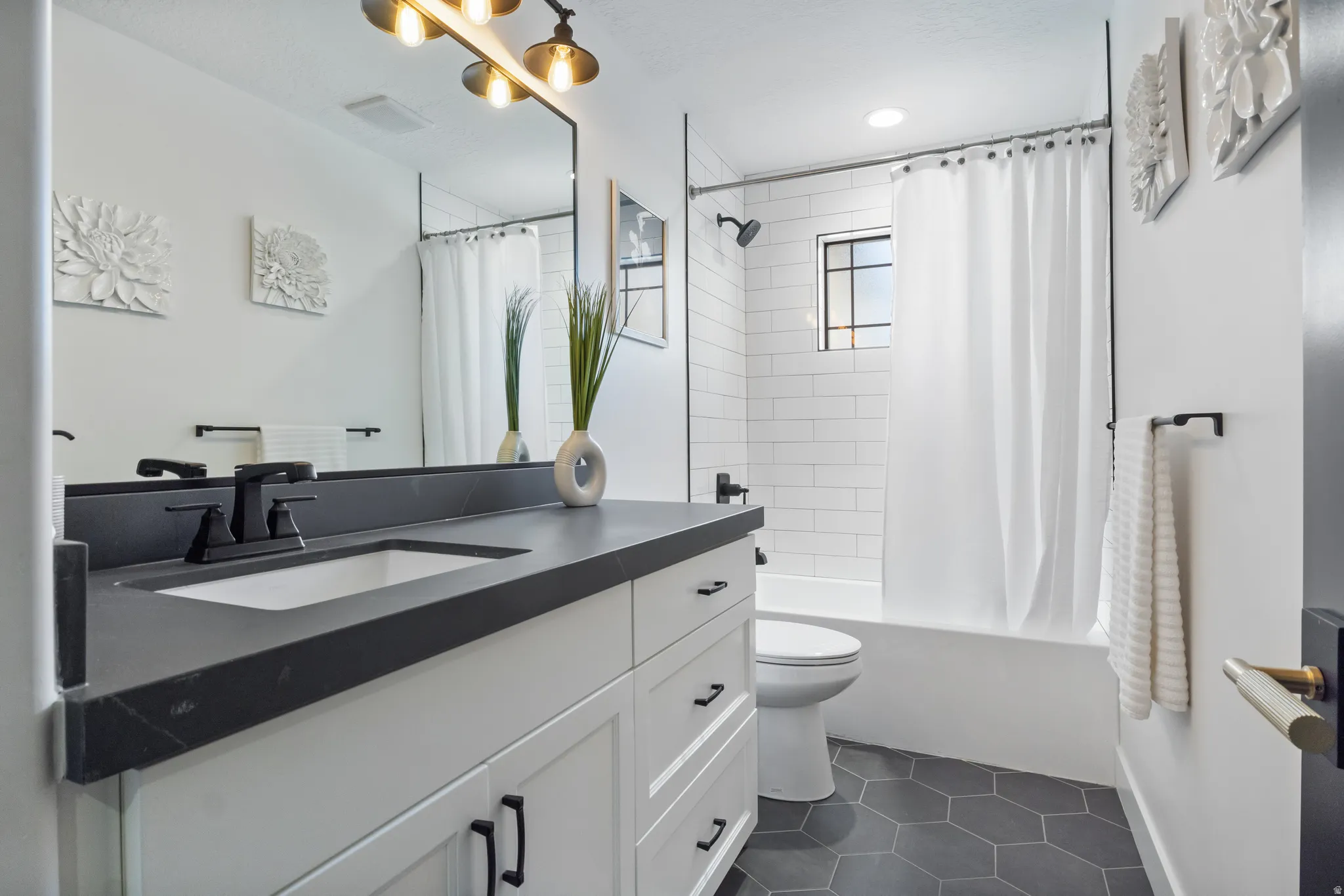 Bathroom featuring a white vanity with a dark countertop and undermount sink, black hardware, a shower-over-tub with subway tile surround, a dark hex-tile floor, and a window