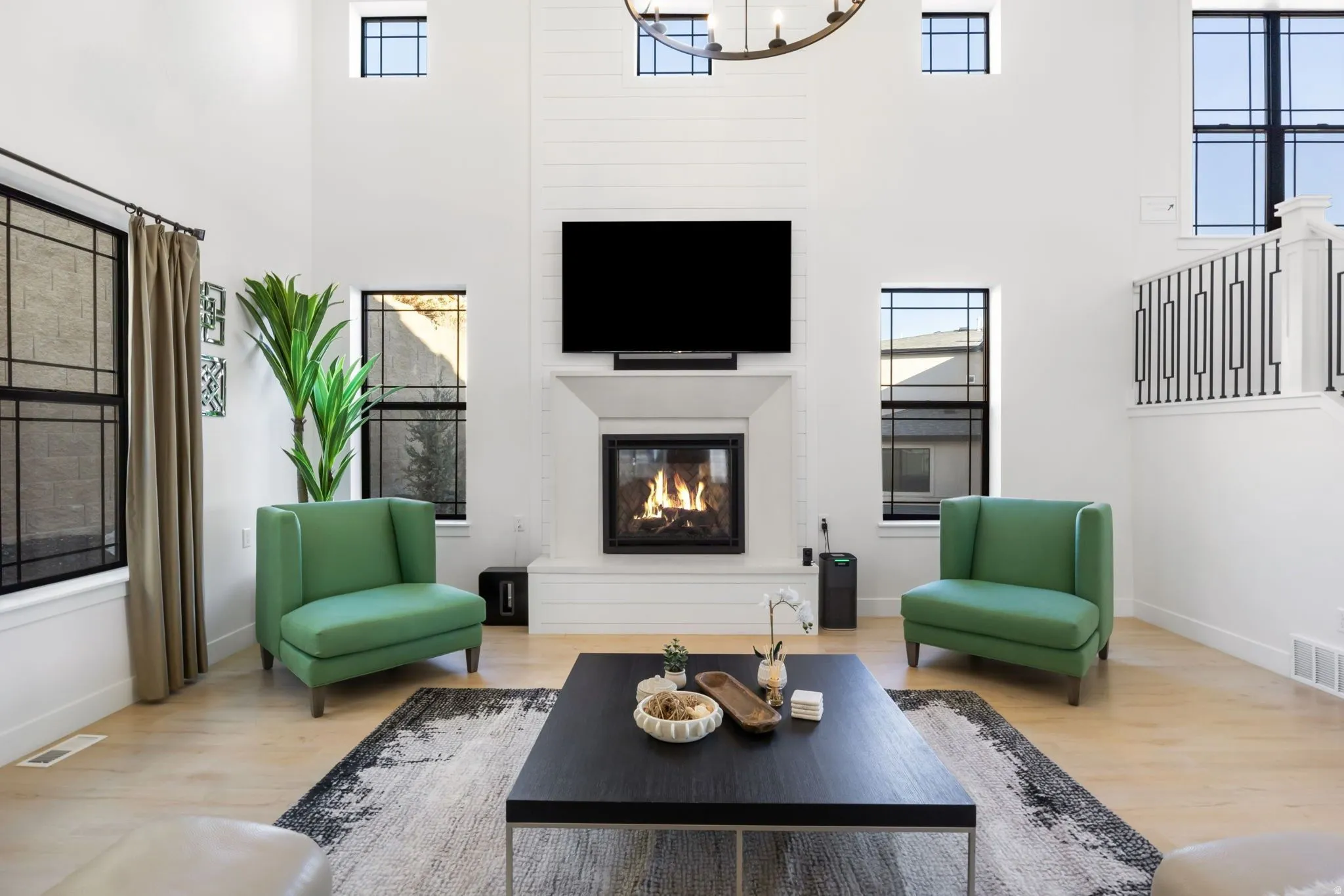 Two-story living area featuring a central fireplace with shiplap accent wall, multiple large windows, light wood-finish flooring, and a contemporary open-tread staircase with metal balusters