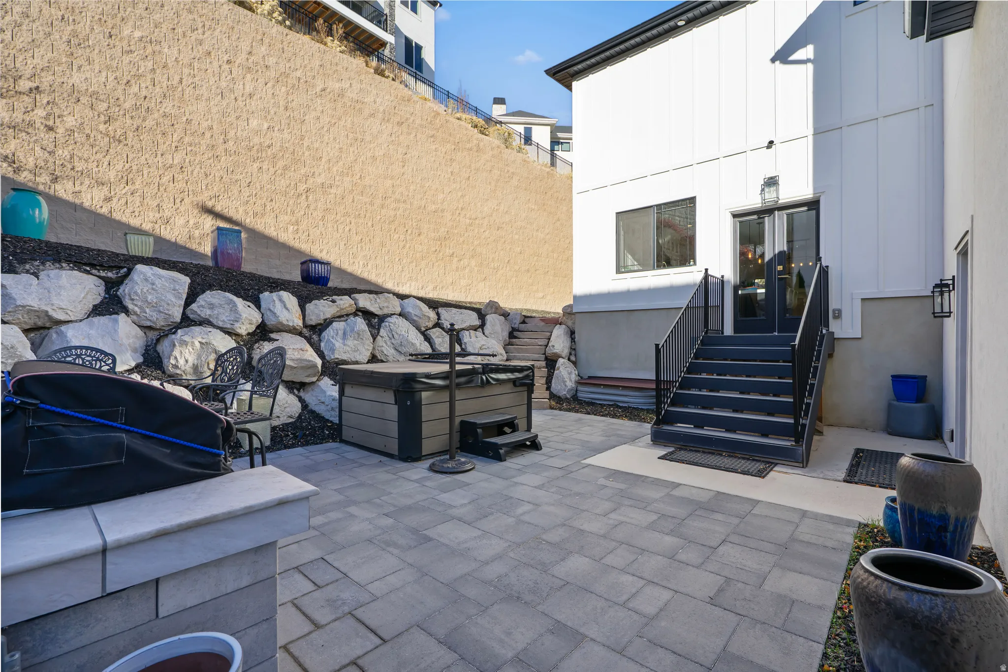 Paved patio area featuring a built-in spa, rock retaining wall, and exterior staircase with black railings leading to a glass-paneled door