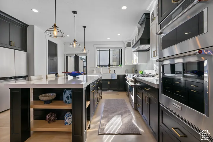 Spacious kitchen featuring a large central island with a light-toned countertop, dark cabinetry, stainless steel appliances, pendant lighting, and light wood-finish flooring