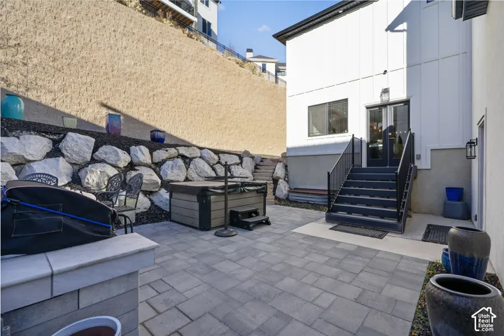 Paved patio area featuring a built-in spa, rock retaining wall, and exterior staircase with black railings leading to a glass-paneled door