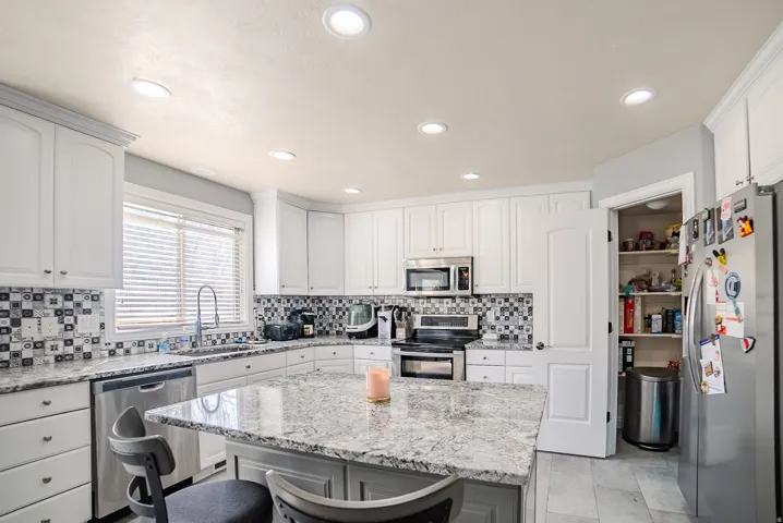 Kitchen featuring an island with a light-toned countertop, white cabinetry with crown molding, stainless steel appliances, recessed ceiling lights, and a tiled backsplash