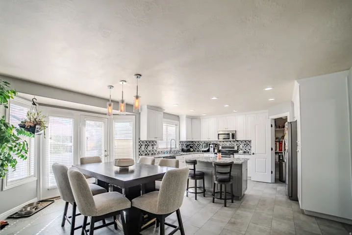 Open-concept kitchen and dining area featuring stainless steel appliances, white cabinetry with contrasting black island, ceramic tile flooring, pendant lighting, and abundant natural light from multi-pane French doors and windows