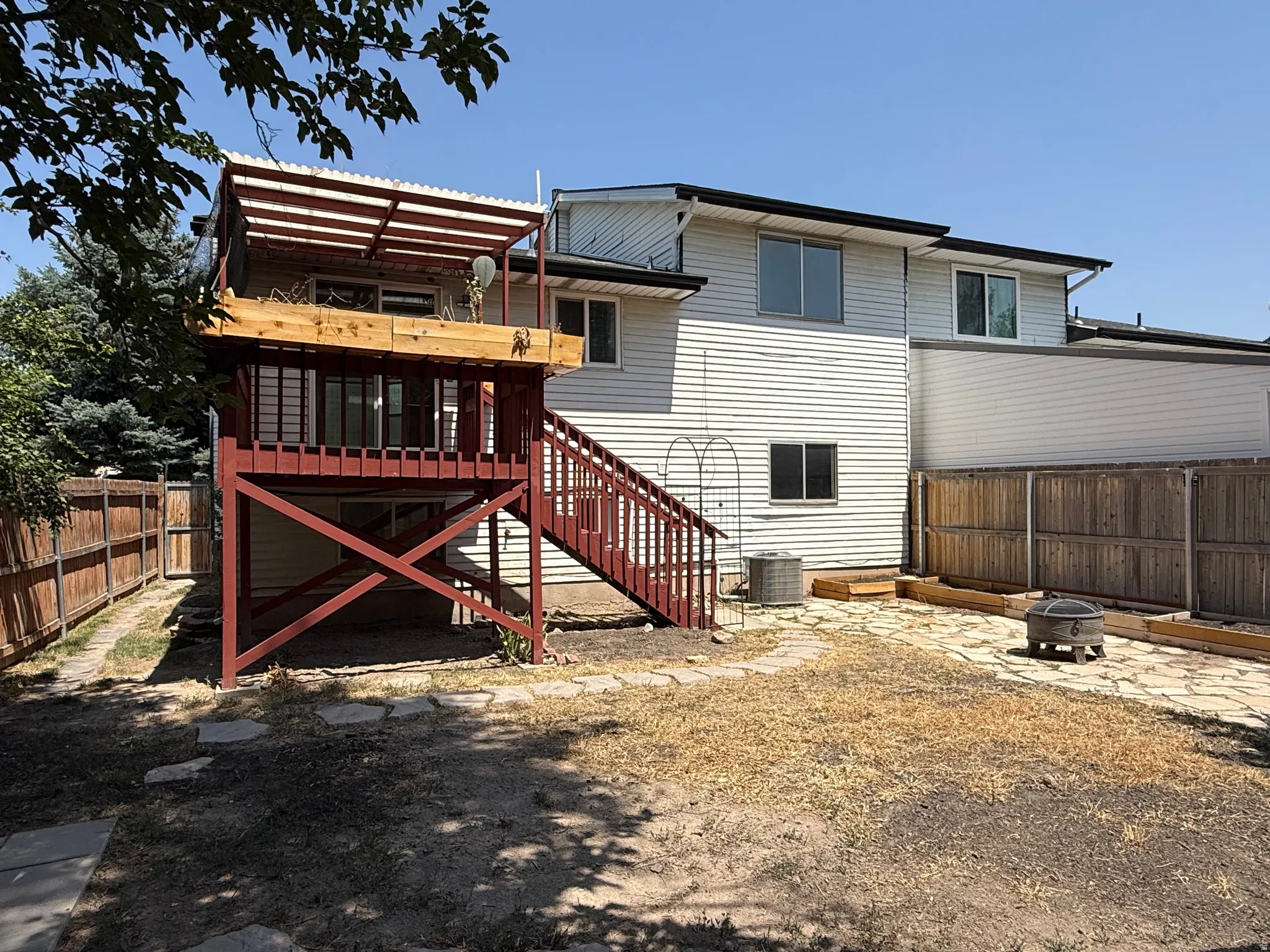 Rear exterior featuring white horizontal siding, a two-story red-stained wood deck with stairs, an upper deck with a corrugated roof, and a fenced yard with a stone patio