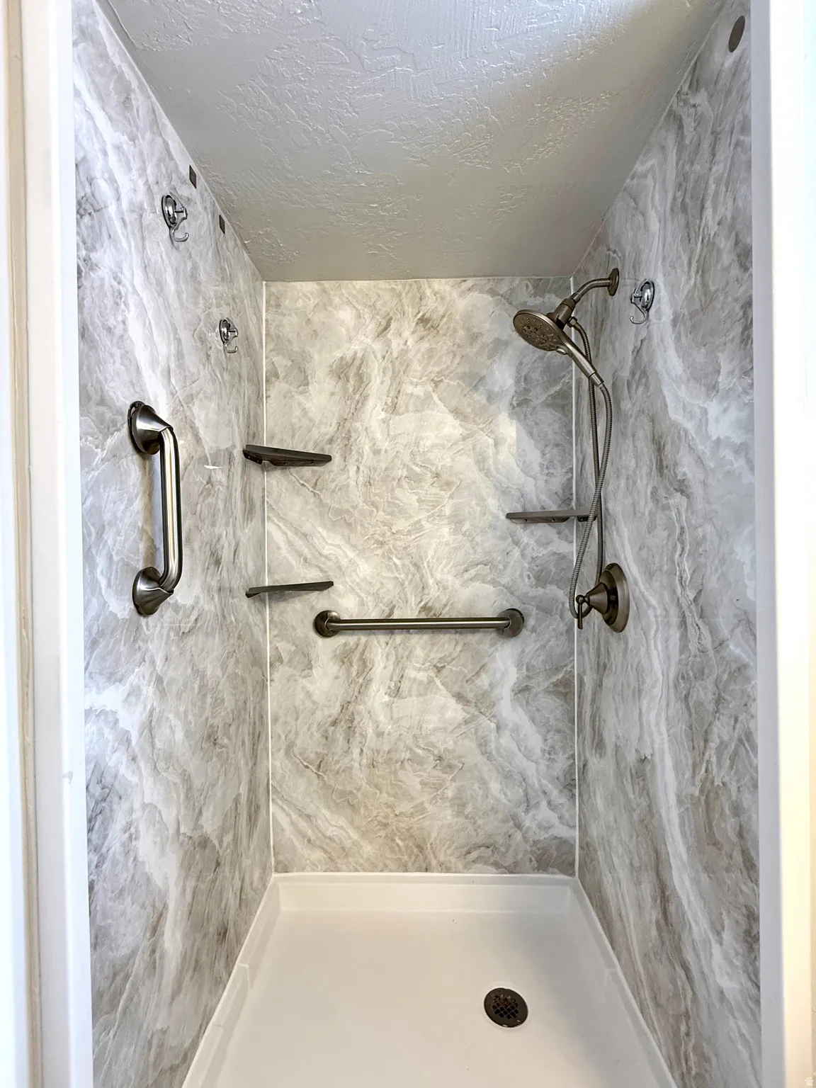 Shower stall with marbled-patterned wall panels, a white shower pan, and a bronze-finish shower head with handheld sprayer