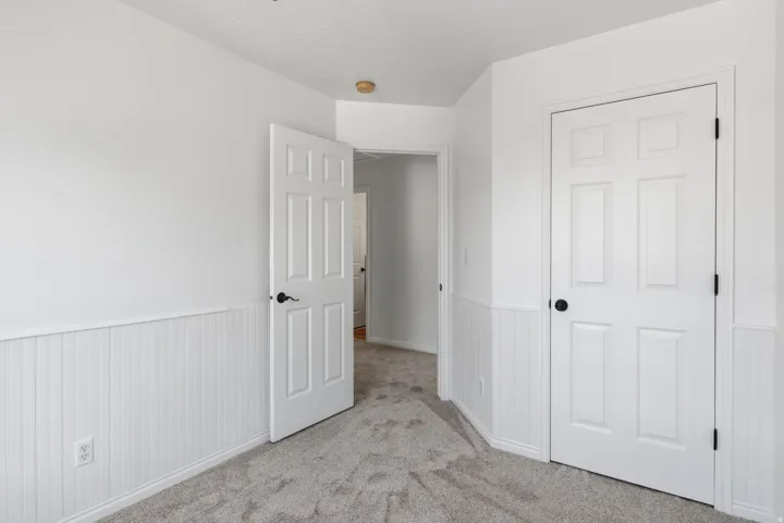 Interior space featuring white walls with wainscoting, textured carpet flooring, and a six-panel door with black hardware