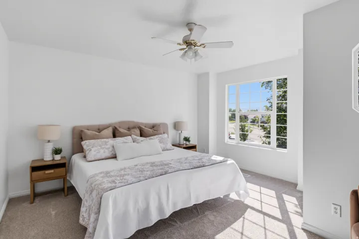 Bright bedroom featuring a ceiling fan, wood-finish nightstands, carpet flooring, and abundant natural light from large windows with exterior visibility