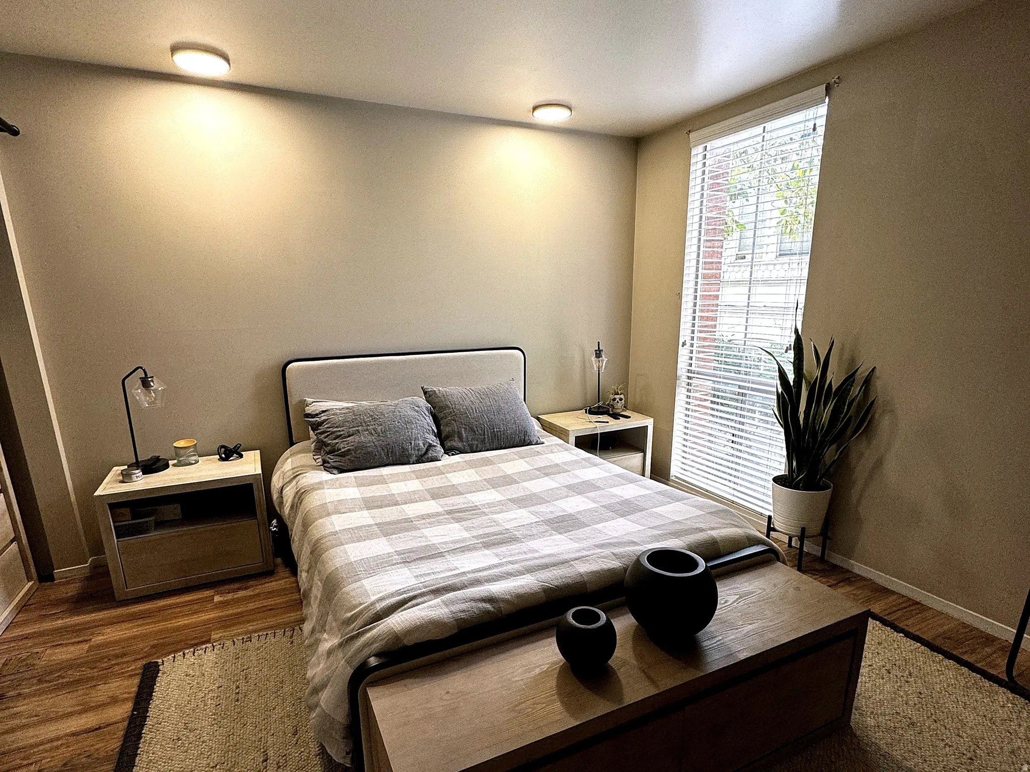 Bedroom featuring wood-finish flooring, a full-height window with horizontal blinds, built-in ceiling lighting, and a neutral wall palette
