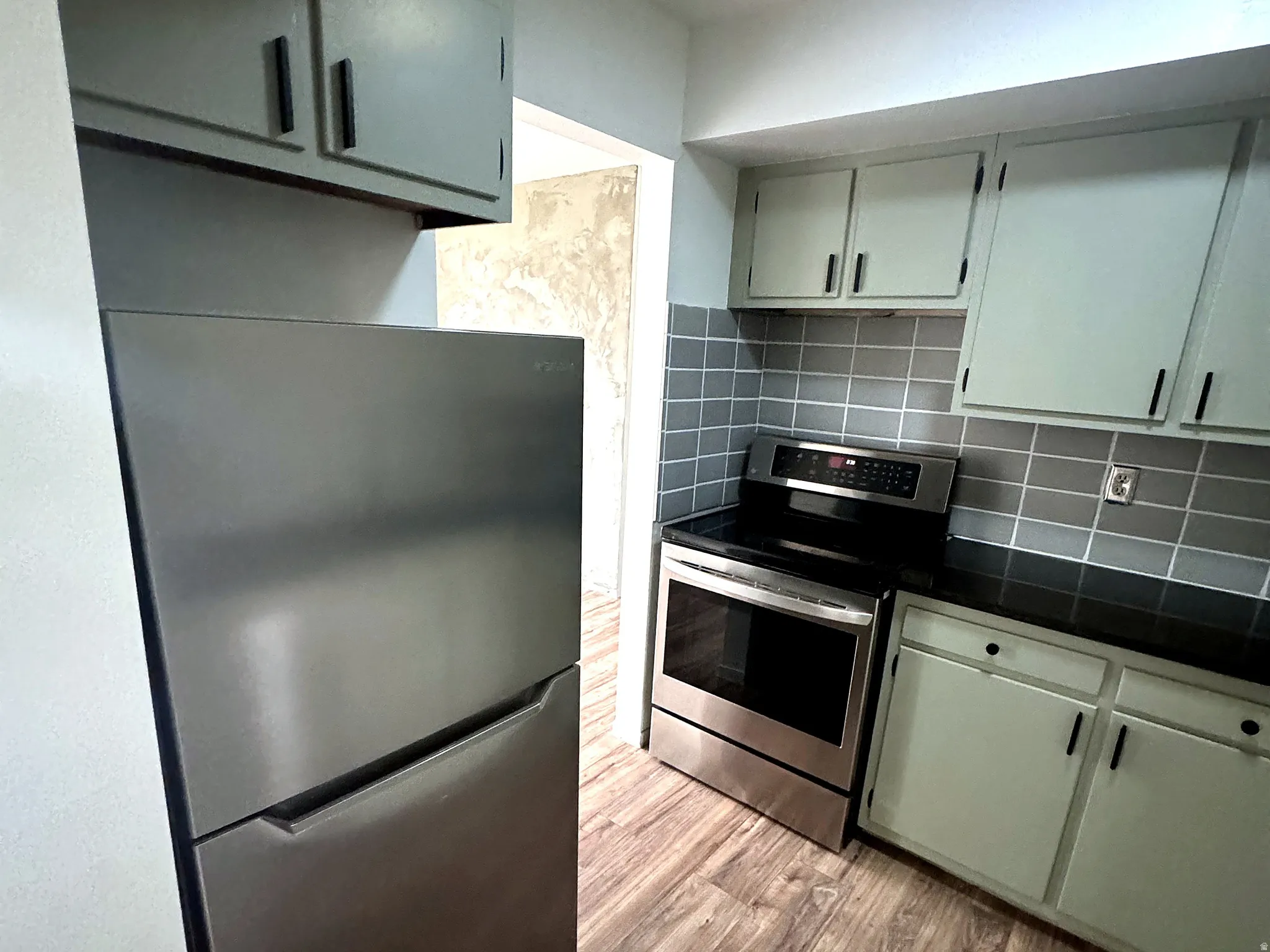 Kitchen featuring stainless steel appliances, dark wood-finish flooring, light-toned cabinetry with dark hardware, black countertops, and a gray tiled backsplash