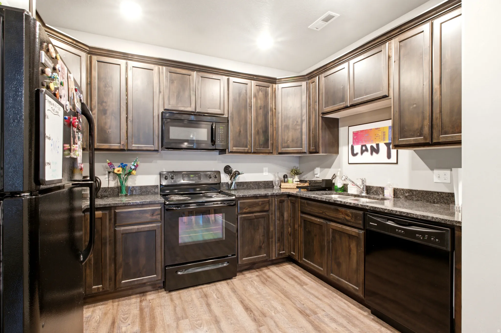Kitchen featuring L-shaped dark wood cabinetry, black appliances, dark speckled countertops, and wood-finish flooring