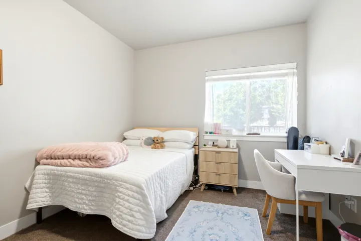 Carpeted bedroom featuring a large window with privacy coverings, light-toned walls, and a built-in desk area