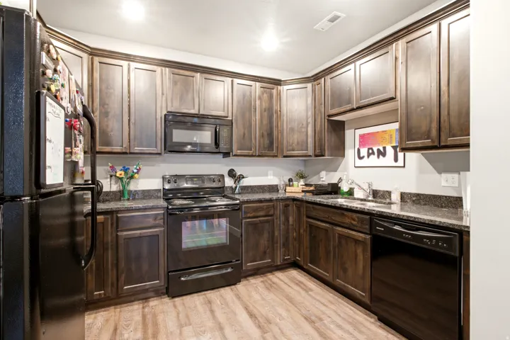 Kitchen featuring L-shaped dark wood cabinetry, black appliances, dark speckled countertops, and wood-finish flooring