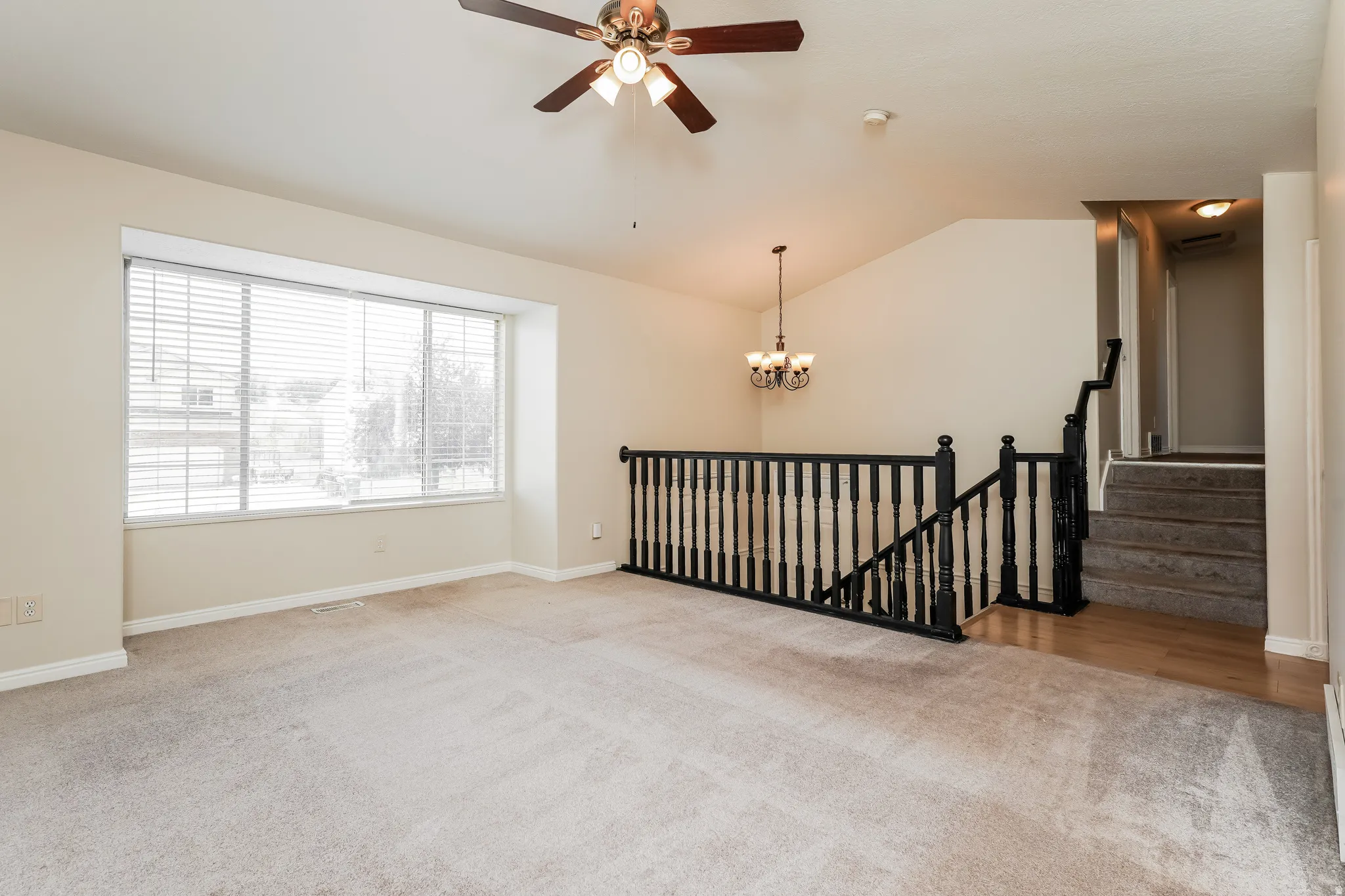 Carpeted living area with vaulted ceiling, large picture window with blinds, and a ceiling fan with integrated lighting
