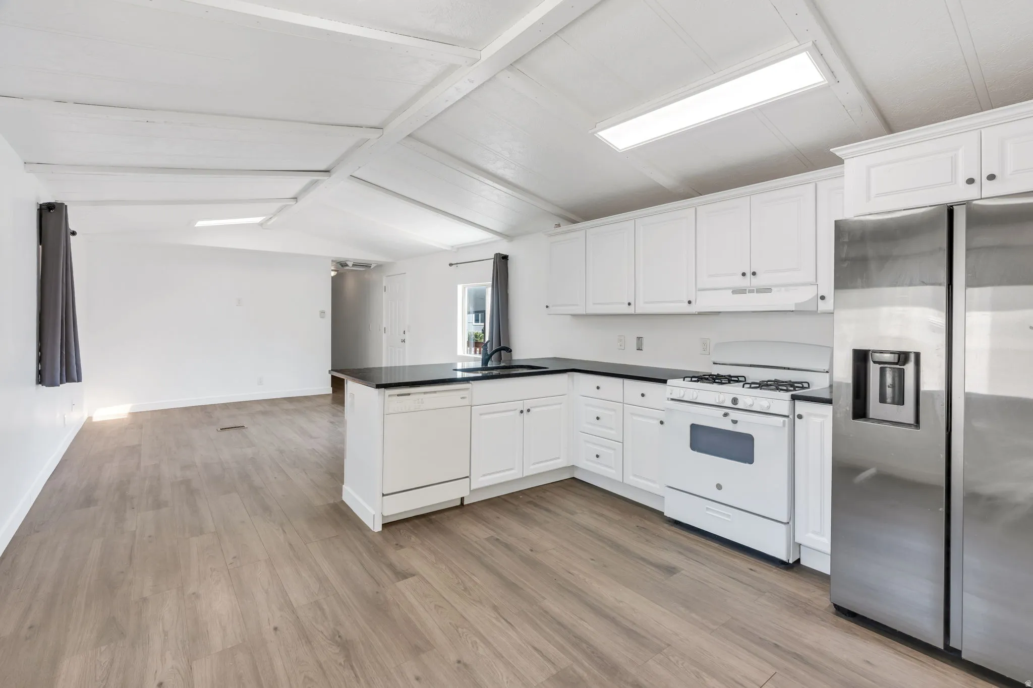 Vaulted ceiling with exposed beams, wood-finish flooring, white cabinetry, dark countertops, and stainless steel refrigerator