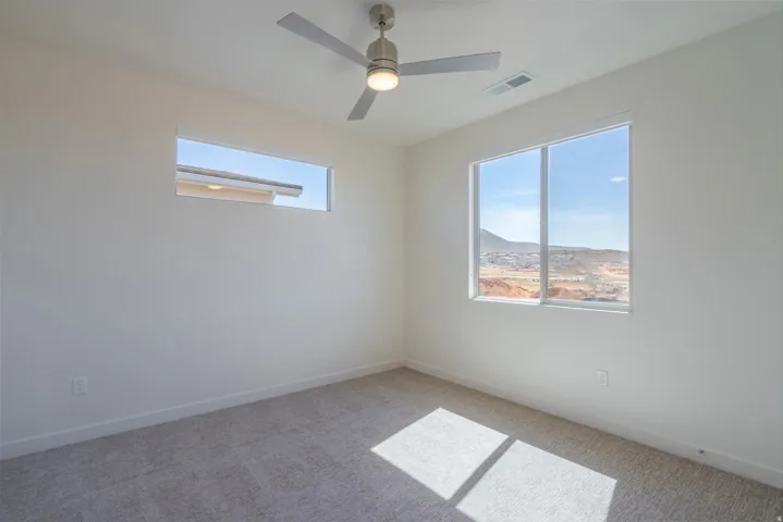 Bright secondary bedroom featuring expansive windows with scenic desert landscape vistas, neutral carpet flooring, ceiling fan with integrated lighting fixture, and abundant natural illumination from dual window openings
