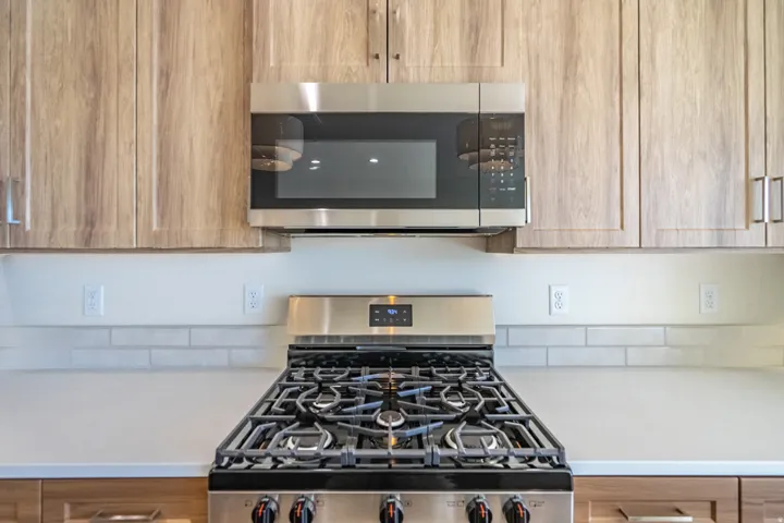 Kitchen featuring a stainless steel range with gas burners, coordinating stainless steel microwave, light wood-finish cabinetry, light-toned countertops, and a white subway tile backsplash