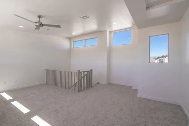Spacious carpeted room featuring a contemporary ceiling fan, recessed lighting, multiple windows, and a wood-finish railing