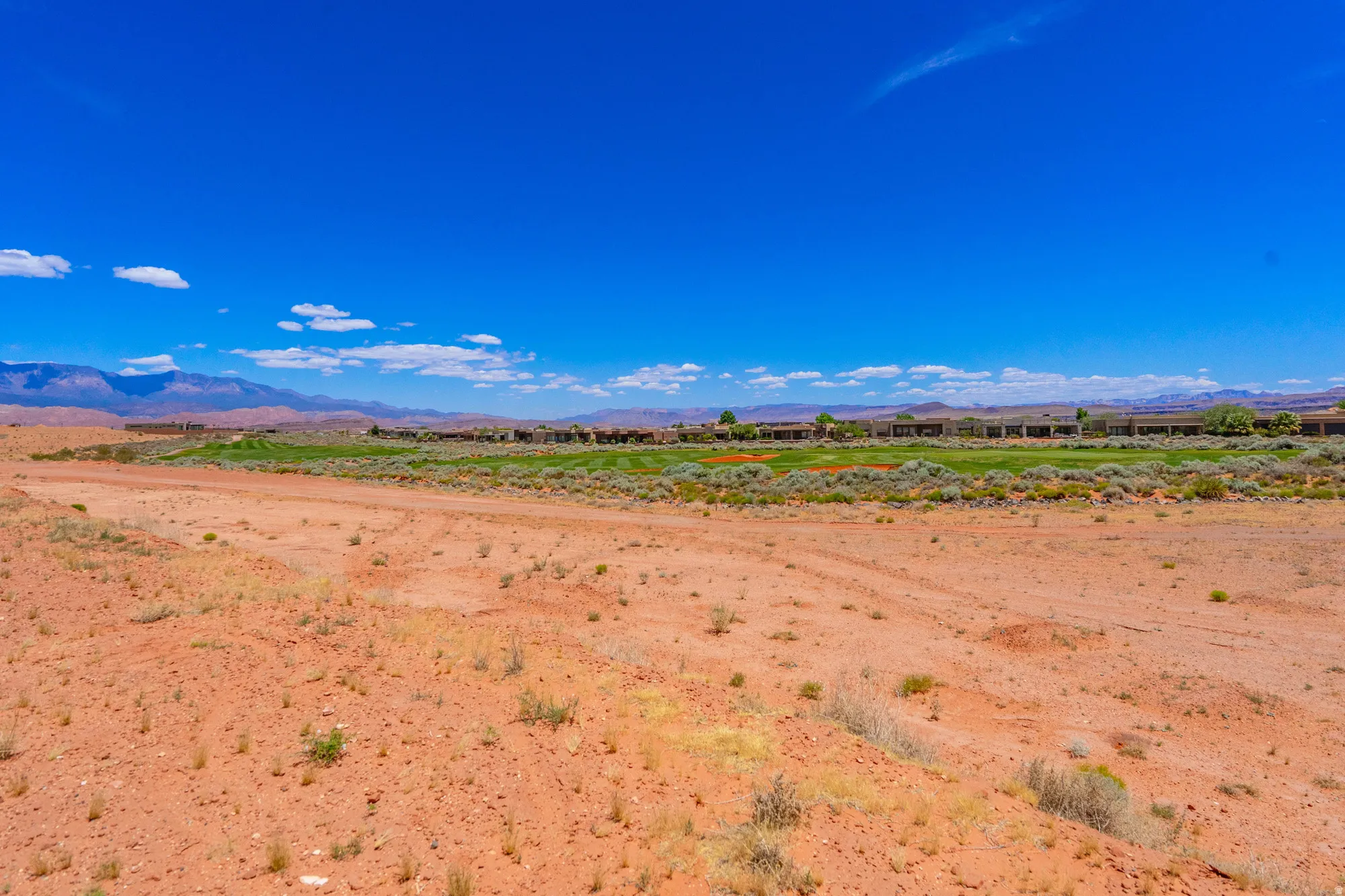 Expansive desert landscape featuring red earth, distant green foliage, and mountain ranges under a clear blue sky