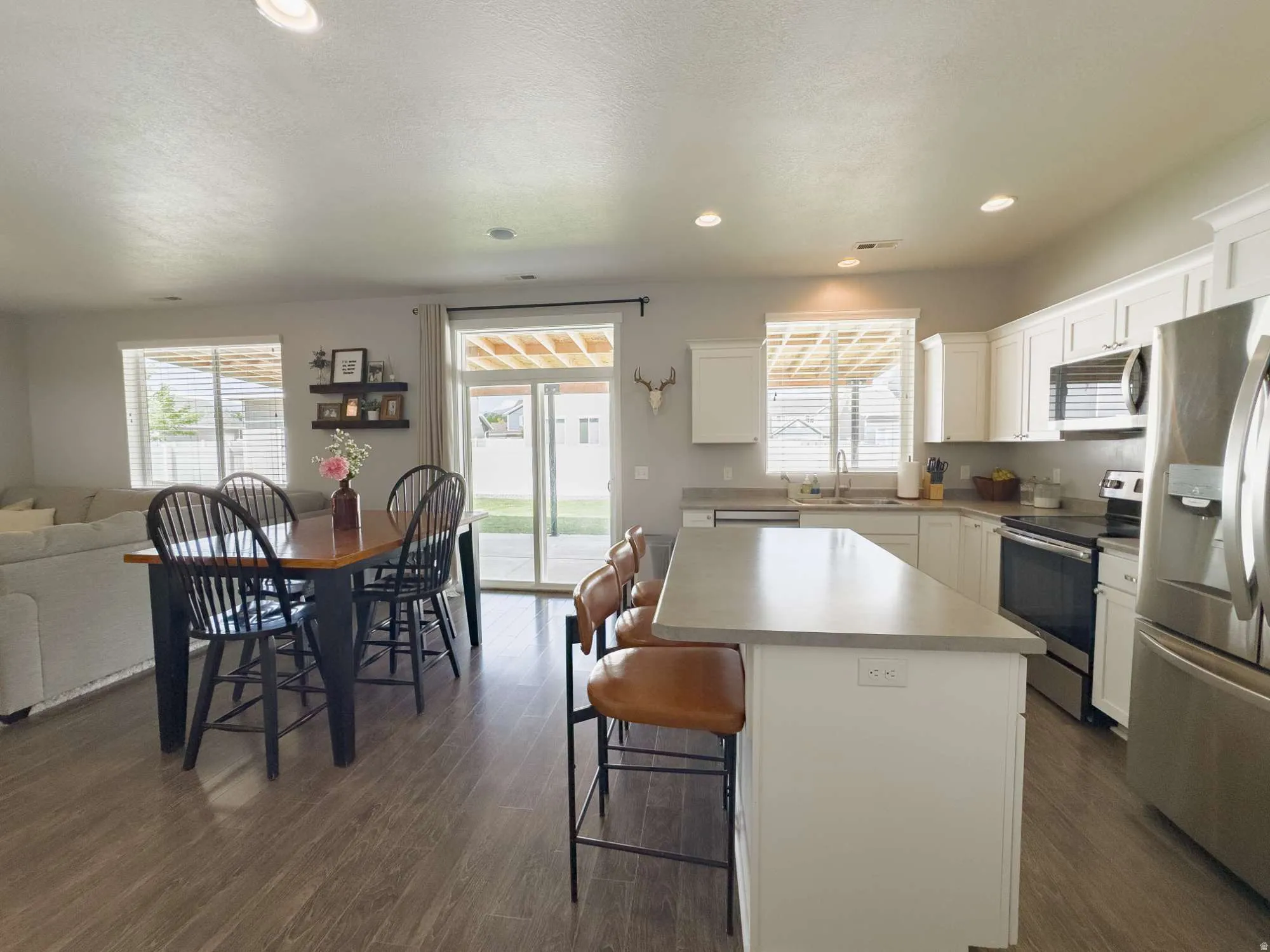 Open-concept kitchen and dining area featuring white cabinetry, stainless steel appliances, a large center island with seating for multiple guests, wood-finish flooring throughout, recessed ceiling lighting, and glass sliding doors providing natural light