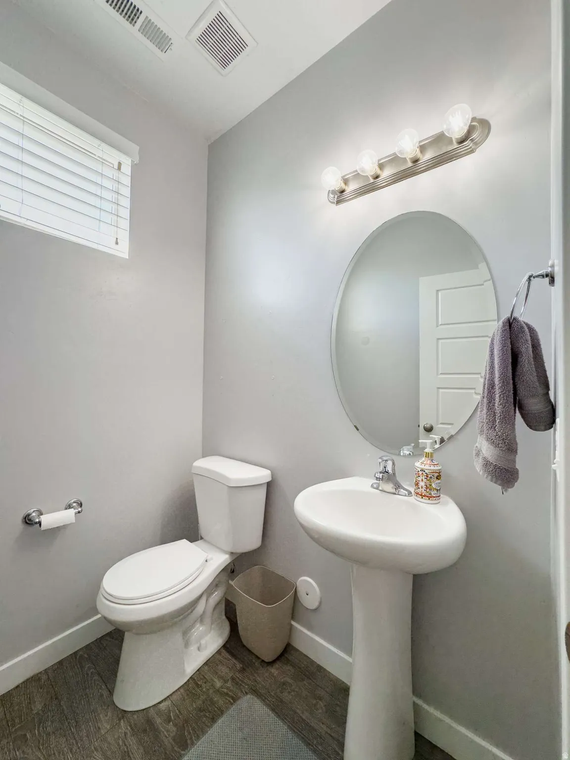 Elegant half bath featuring a pedestal sink with chrome fixtures, large oval mirror with integrated lighting, and neutral gray wall finishes