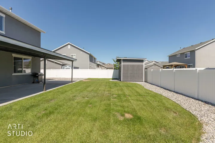 Expansive lawn with a covered concrete patio, gravel border, private vinyl fencing, and an exterior storage shed
