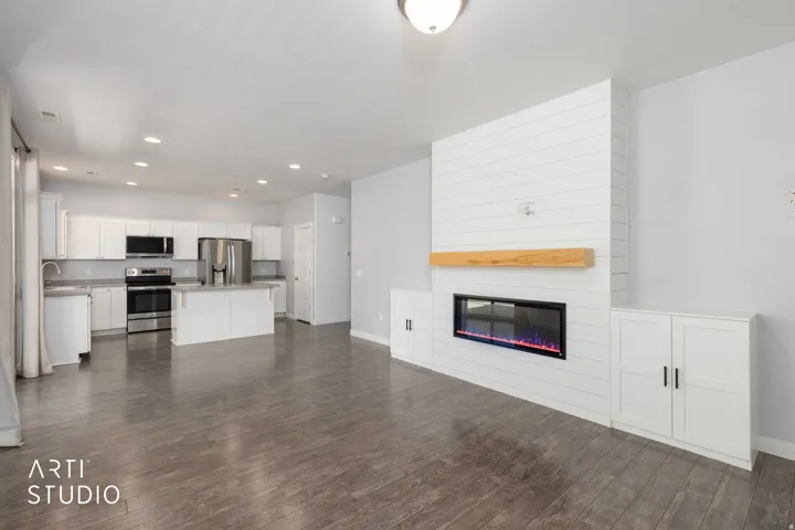 Open-concept living area featuring wood-finish flooring, a contemporary fireplace with a wood mantle, white built-in cabinetry, recessed lighting, and a kitchen with stainless steel appliances