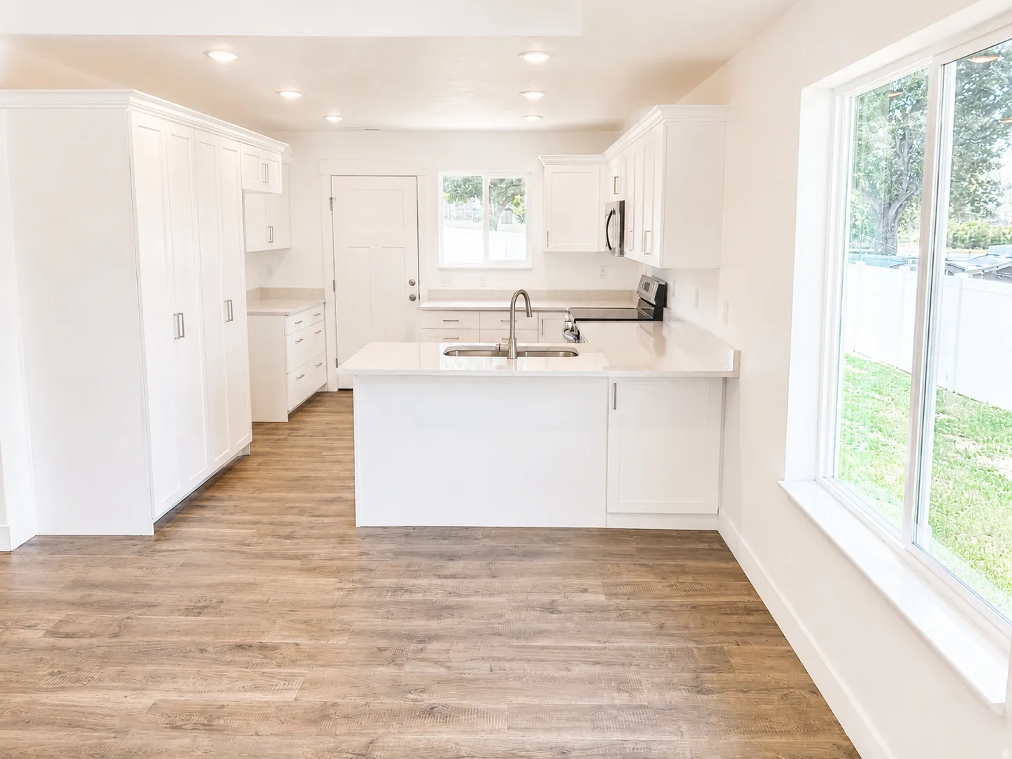 Open-concept kitchen featuring white shaker cabinetry, an island with an undermount sink, light-toned countertops, integrated stainless steel appliances, and wood-finish flooring