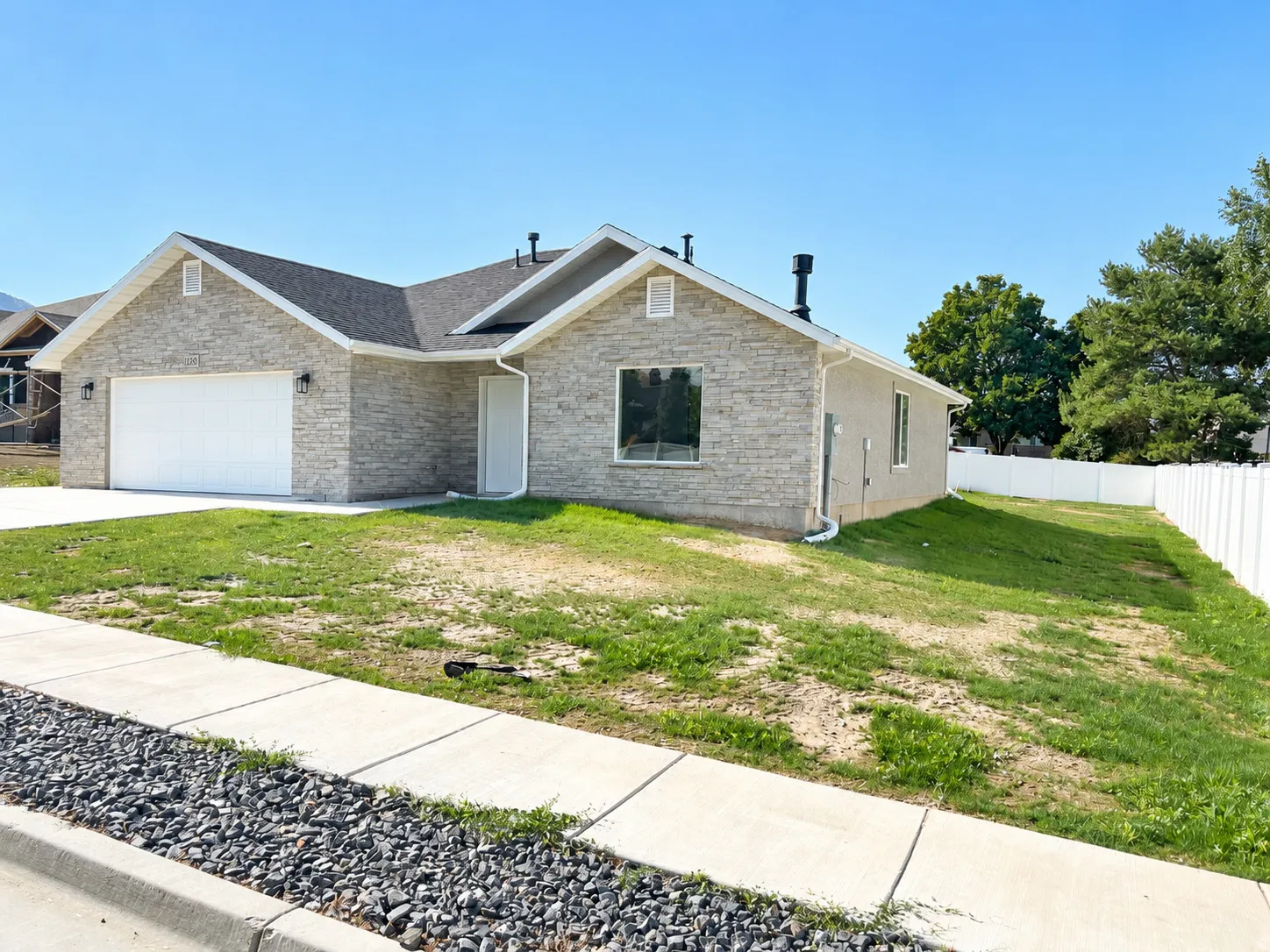 Stone facade home featuring a two-car garage, white vinyl fencing, a concrete driveway, and a sidewalk with a rock-lined strip