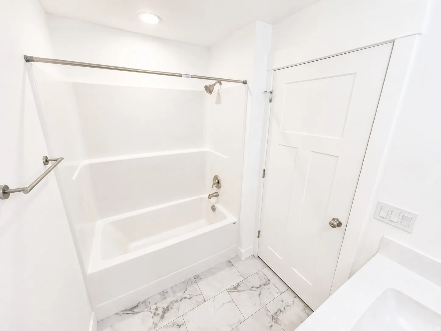 Bathroom featuring a white tub and shower combination with brushed nickel fixtures, complemented by a three-panel interior door with a brushed nickel knob, white quartz countertop, and white marble-look tile flooring