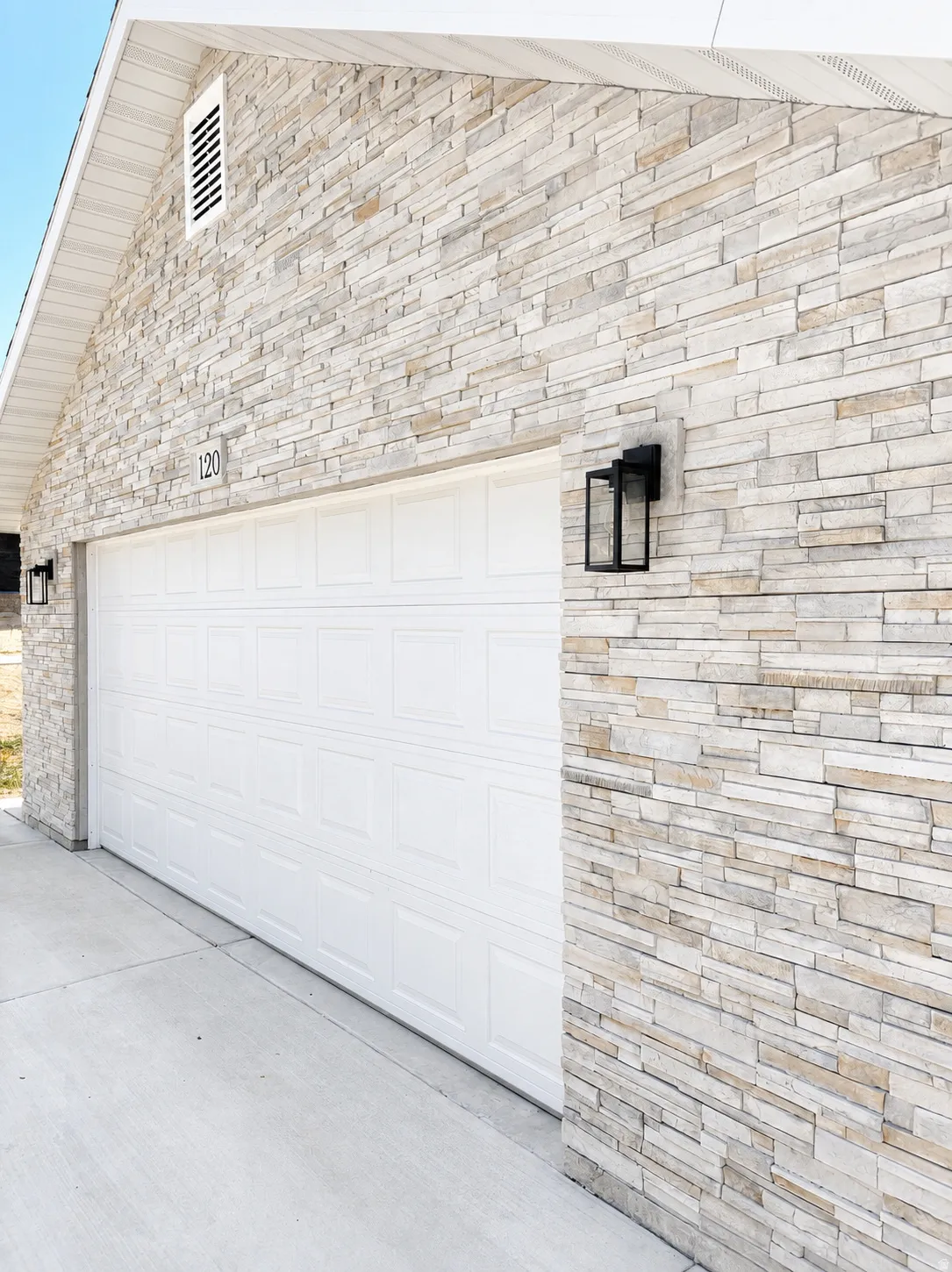 Stone veneer exterior featuring a paneled garage door, modern black wall sconces, white soffit, and a concrete driveway