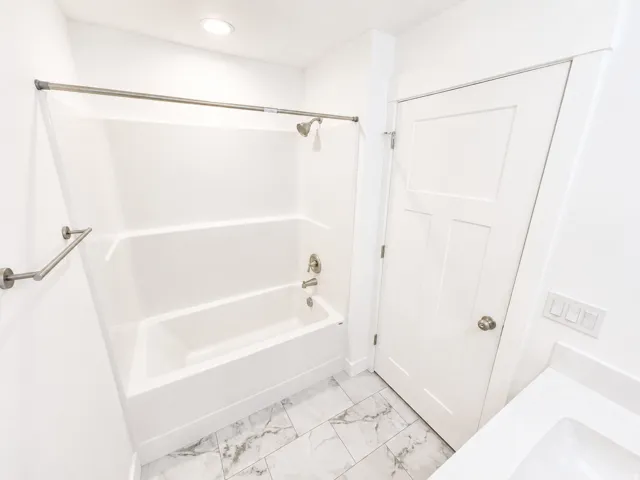 Bathroom featuring a white tub and shower combination with brushed nickel fixtures, complemented by a three-panel interior door with a brushed nickel knob, white quartz countertop, and white marble-look tile flooring