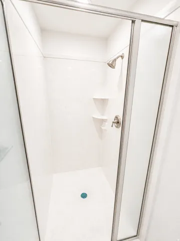 Master shower enclosure featuring a brushed nickel showerhead and faucet, white paneling, integrated corner shelving, and a clear glass sliding door with a brushed nickel frame