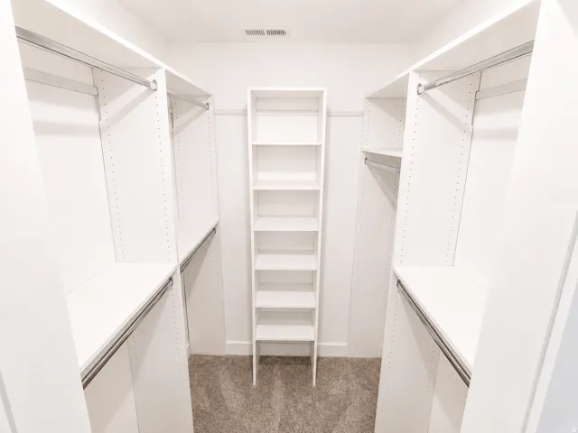 Master Walk-in closet featuring white built-in shelving, multiple hanging rods, and a central freestanding shelving unit over carpeted flooring