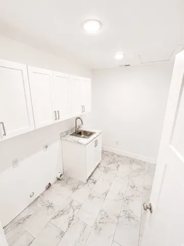 Utility room featuring white shaker cabinetry, a stainless steel sink with a gooseneck faucet, a white countertop with a gray veining pattern, and light-toned rectangular floor tiles