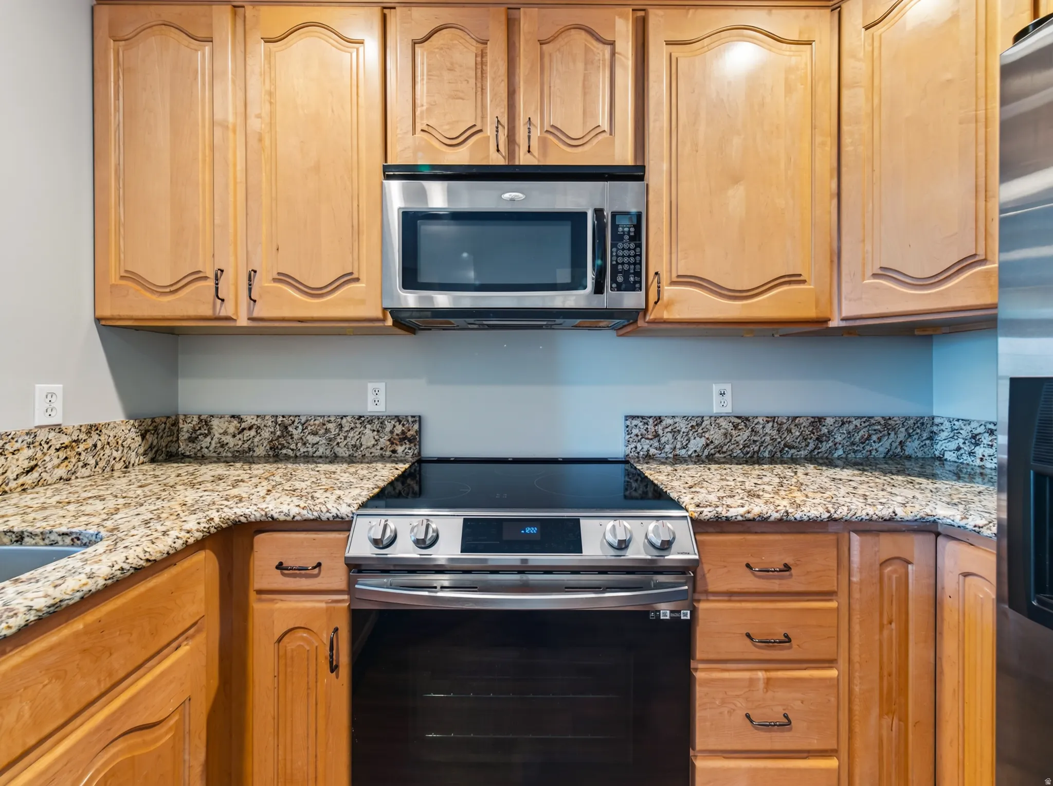 Kitchen featuring light wood cabinetry with arched panel doors, stone-finish countertops, stainless steel microwave, and a black glass-top range