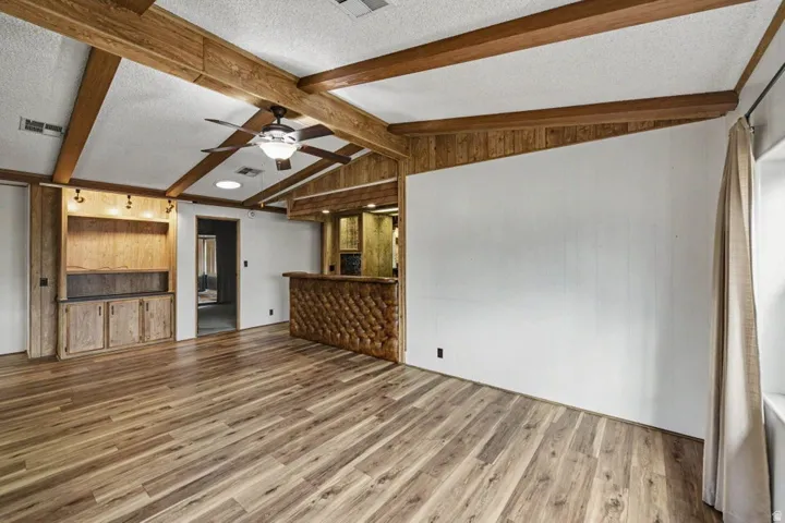 Spacious living area featuring wood-finish flooring, exposed ceiling beams, a built-in entertainment center with integrated lighting, a ceiling fan, and a built-in bar with a textured front