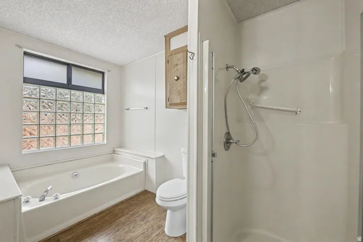 Bathroom featuring a full-size tub, glass block window, wood-finish flooring, a separate shower stall with a grab bar, and a wall-mounted wooden cabinet