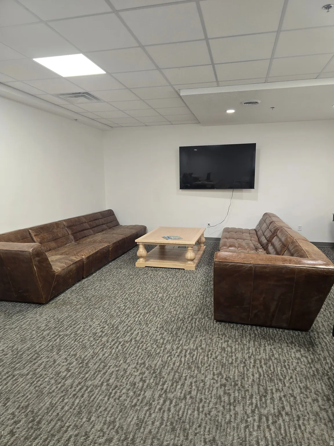 Spacious room with patterned carpet flooring, drop ceiling with integrated lighting, and a wall-mounted flat-screen display