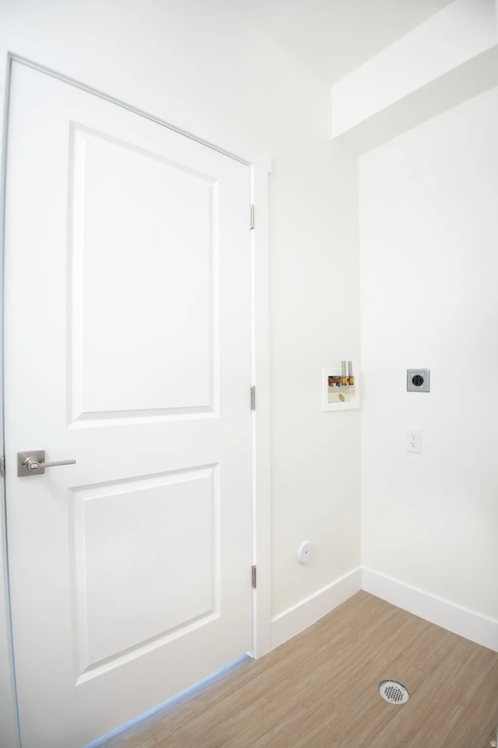 Utility space featuring wood-finish flooring, white paneled door with brushed nickel hardware, crisp white walls, and a floor drain