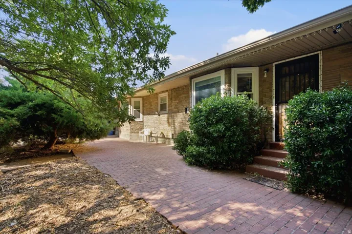 Brick exterior featuring a bay window, a red brick paver walkway, mature trees, and established shrubbery