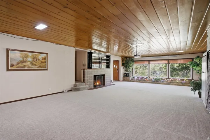 Spacious room featuring a wood-paneled ceiling, light-toned carpet flooring, a tiled fireplace with hearth, and large windows
