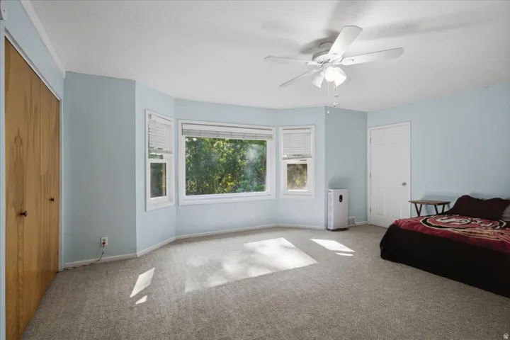 Spacious room featuring light blue walls, gray carpet, a multi-pane bay window, a ceiling fan with integrated lighting, and a wood-finish closet door