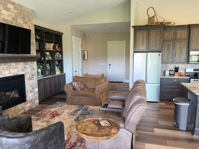 Spacious open-concept interior featuring wood-finish flooring, a stone-faced fireplace with a wood mantle, custom built-in cabinetry, and a kitchen with stone-look countertops and matching backsplash