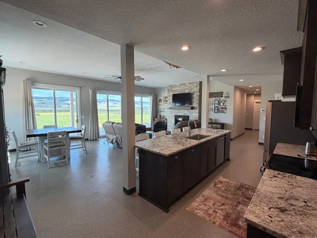 Open-concept living area with extensive sliding glass doors, a stone-faced fireplace, a large kitchen island with integrated sink, granite countertops, and recessed lighting